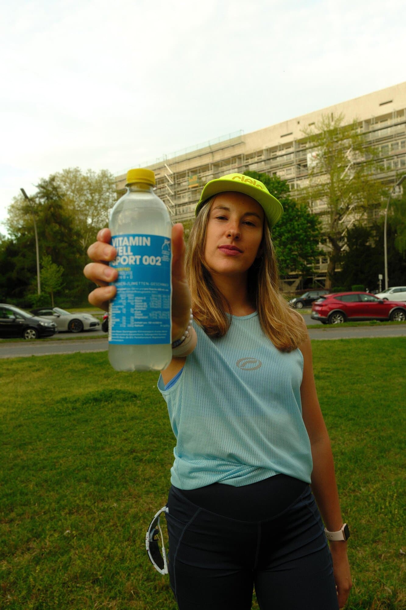 A woman in athletic attire offers a bottle labeled Vitamin Well Sport 002 outdoors.
