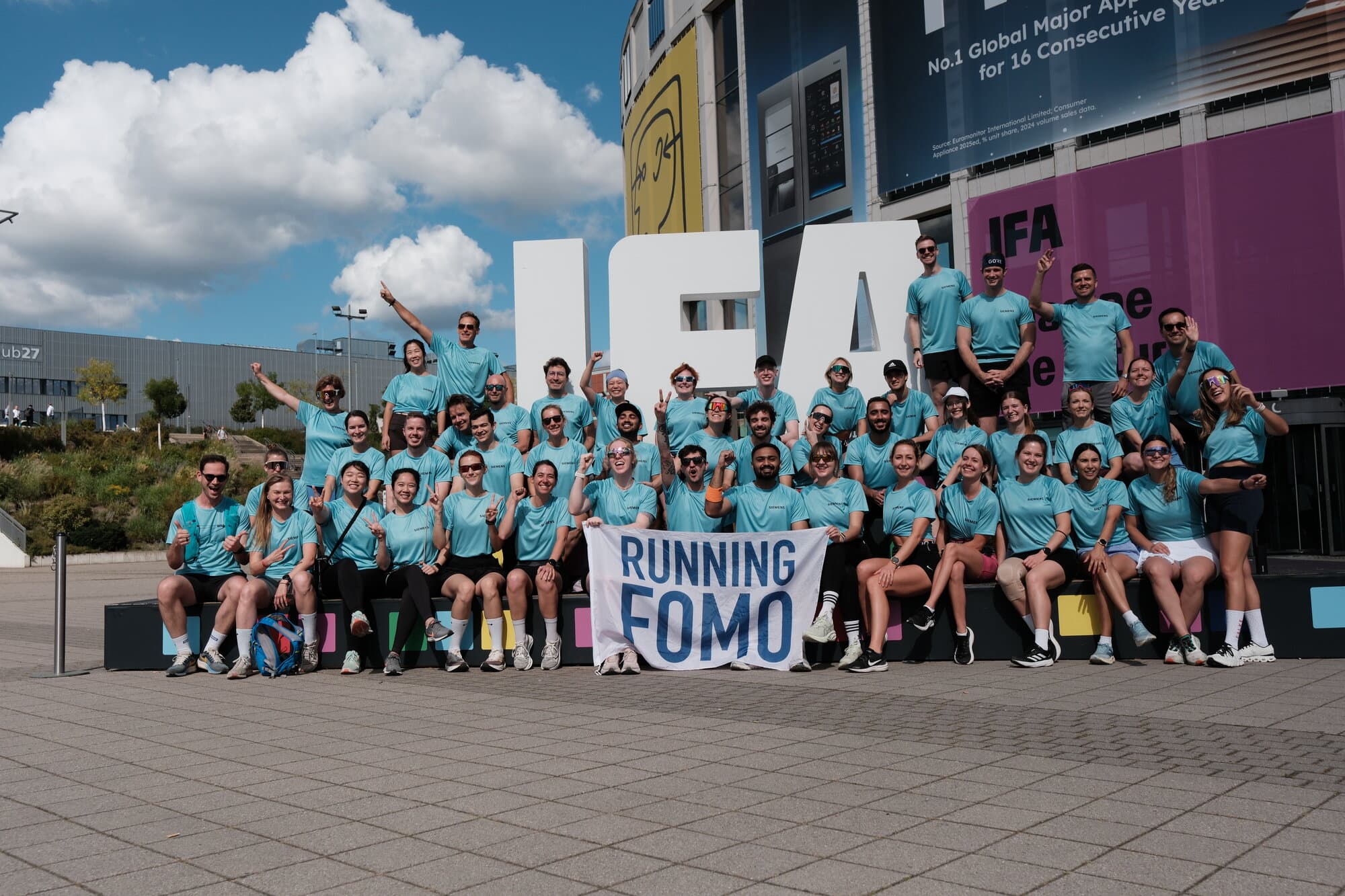 A group of people in matching shirts pose with a Running FOMO banner at an event.