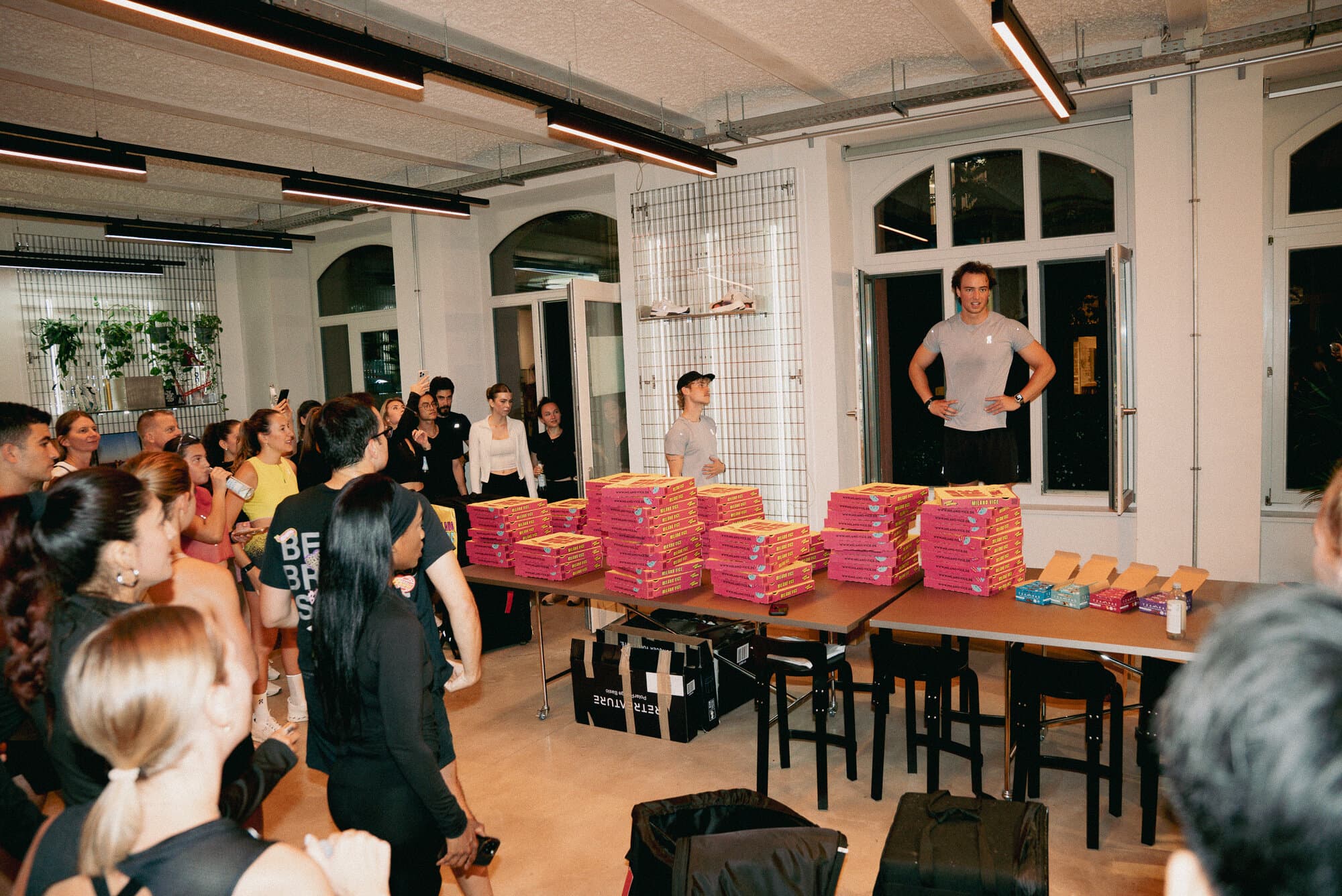 A group gathers around tables stacked with pizza boxes in a well-lit room.